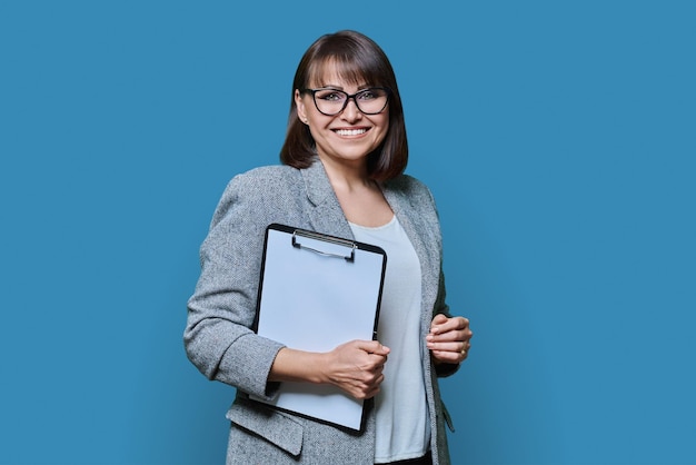Femme d'affaires avec un bloc-notes sur un fond bleu femme d'âge moyen confiante en veste à lunettes regardant la caméra tenant des documents sur le bloc-notices Personnel de direction de la gestion des affaires