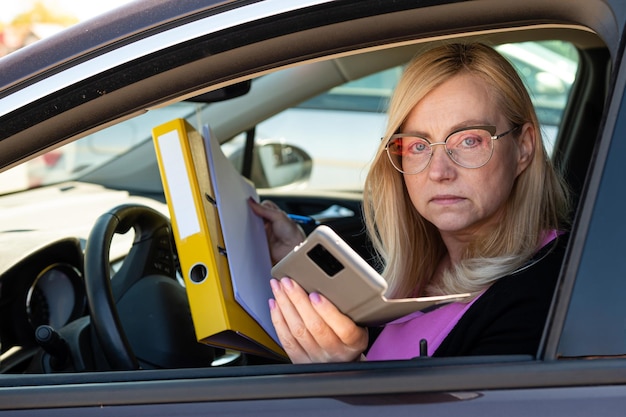 Femme d'affaires blonde d'âge moyen dans des verres en voiture travaillant avec le concept de technologie mobile de documents