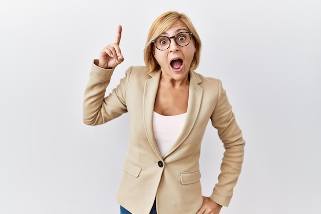 Femme d'affaires blonde d'âge moyen debout sur fond isolé pointant le doigt vers le haut avec une idée réussie sorti et heureux numéro un