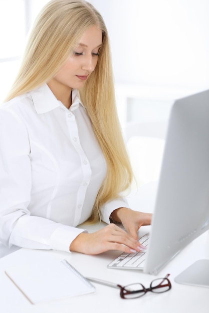 Femme d'affaires blonde assise et travaillant avec un ordinateur dans un bureau blanc