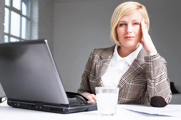 Une femme d'affaires blonde en costume élégant a des maux de tête de travail à l'aide d'une pilule effervescente sur l'eau dans un lieu de travail en verre avec un ordinateur portable dans un bureau blanc