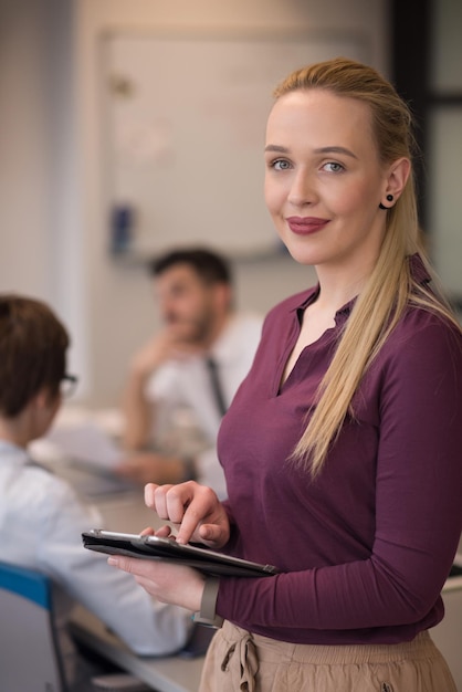 Femme d'affaires blonde dans des vêtements décontractés travaillant sur une tablette à l'intérieur du bureau d'affaires de démarrage moderne. Groupe de jeunes lors d'une réunion d'équipe estompée en arrière-plan