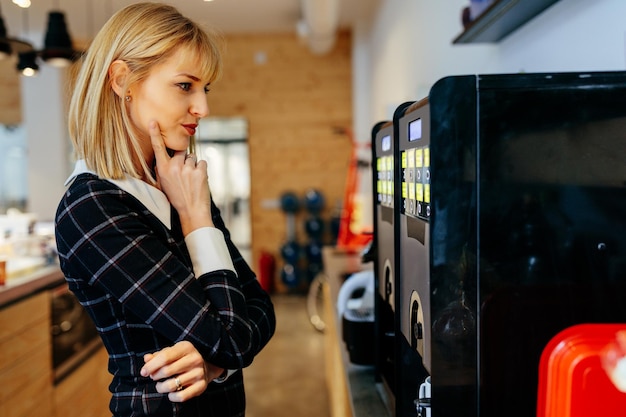 Femme d'affaires blonde debout devant une cafetière dans une cafétéria de bureau
