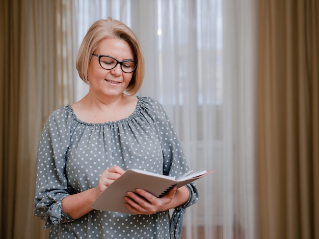 Femme d'affaires blonde, souriante et âgée, portant des lunettes, lisant et naviguant avec un agenda quotidien et un stylo à bille. Portrait d'un beau retraité bien soigné de 50 à 60 ans dans la chambre.