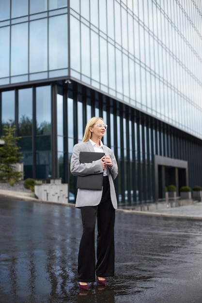 Femme d'affaires blonde tenant un ordinateur portable en détournant les yeux en attendant quelqu'un dans la rue un jour de pluie