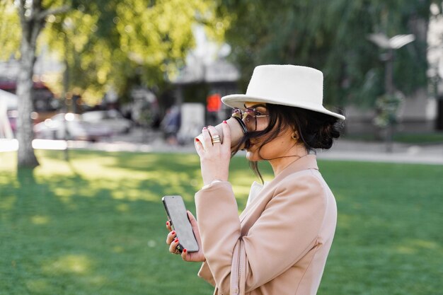 Une femme d'affaires boit du café en plein air dans le parc. La fille portait un chapeau blanc, une robe et une veste.