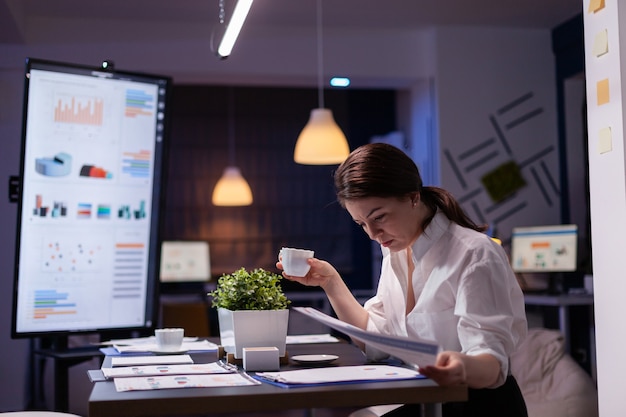 Femme d'affaires bourreau de travail regardant la présentation des rapports sur le moniteur en comparant avec le graphique financier sur la paperasse. Gestionnaire ciblé travaillant dans la salle de réunion d'une entreprise commerciale tard dans la nuit