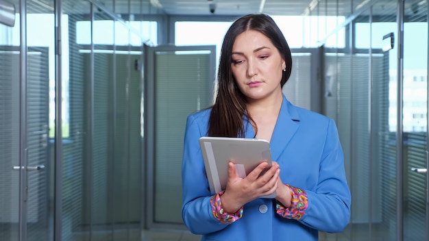 Femme d'affaires brune aux cheveux longs en veste bleue glisse une tablette grise debout dans le salon des locaux parmi les bureaux avec porte en verre transparent, copyspace