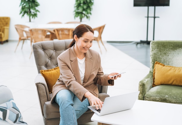 Femme d'affaires brune souriante adulte de quarante ans aux cheveux longs en costume beige élégant et jeans travaillant sur un ordinateur portable au bureau de l'espace ouvert de la place publique