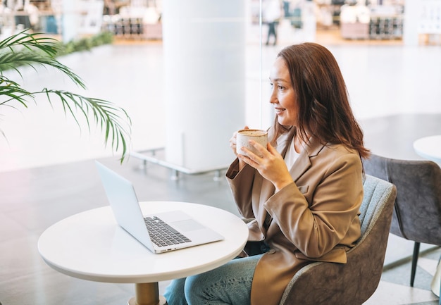 Femme d'affaires brune souriante adulte de quarante ans aux cheveux longs en costume beige élégant travaillant sur un ordinateur portable en buvant du café au café