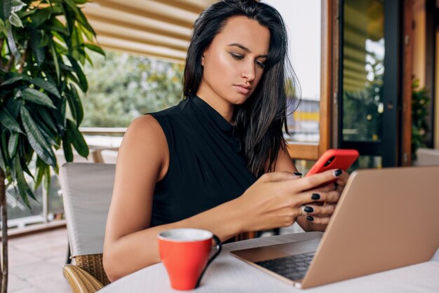 Femme d'affaires brune travaillant sur un ordinateur portable à l'intérieur dans une cafétéria Image de jeunes femmes sérieuses envoyant des SMS sur un téléphone portable avec du café dans un restaurant