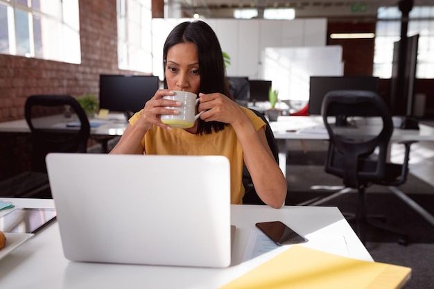 Femme d'affaires caucasienne assise au bureau à l'aide d'un ordinateur portable et buvant une tasse de café