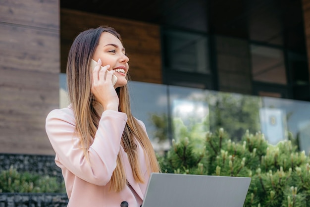 Femme d'affaires caucasienne parlant au téléphone près de l'immeuble de bureaux avec un ordinateur portable dans les mains