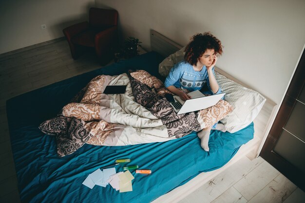 Femme d'affaires caucasienne réfléchie aux cheveux bouclés assis sur le canapé avec un ordinateur portable en prenant quelques notes dans le livre