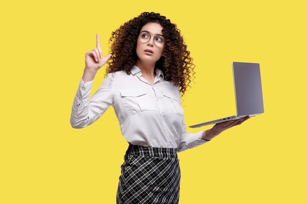 Femme d'affaires caucasienne surprise en chemise blanche et lunettes avec ordinateur portable et pointant vers le haut le doigt sur le mur isolé jaune.