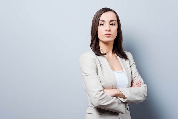 Femme d'affaires charmante. Jeune femme d'affaires confiante gardant les bras croisés et regardant la caméra en se tenant debout sur fond gris