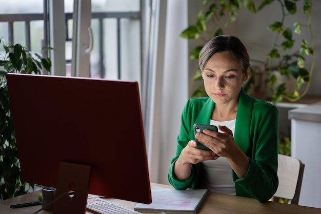 Une femme d'affaires ciblée tenant un téléphone portable est assise dans un bureau moderne à table avec un ordinateur