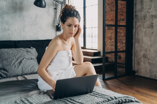 Femme d'affaires ciblée travaille sur un ordinateur portable assis sur le lit après la douche le matin à la maison Travail à distance