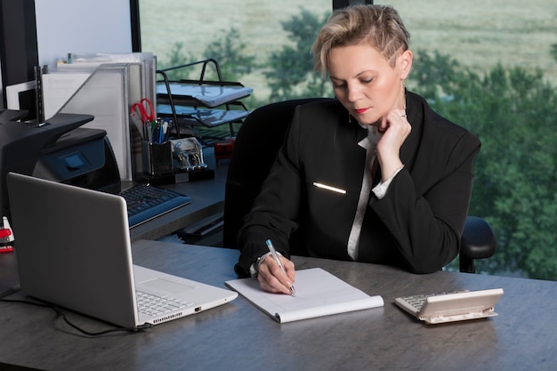 Une femme d'affaires comptable en costume noir au bureau utilise une calculatrice et écrit des données dans un cahier. Analyse des bénéfices, calculs d'impôts et de paiements, préparation du concept d'états financiers