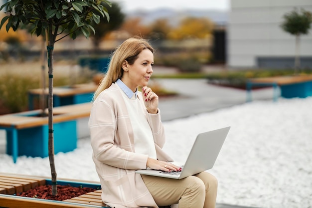 Une femme d'affaires concentrée pense à faire un rapport alors qu'elle est assise dans un parc du centre-ville avec un ordinateur portable