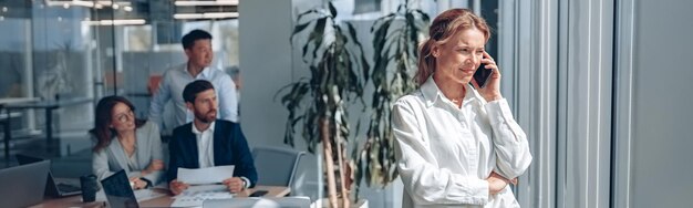 Une femme d'affaires concentrée se tient près de la fenêtre et parle au téléphone pendant une réunion dans un bureau moderne