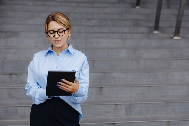 Femme d'affaires confiante aux cheveux blonds permanent office center et travaillant sur tablette numérique. Jeune femme à lunettes utilisant des gadgets modernes pour travailler à distance.