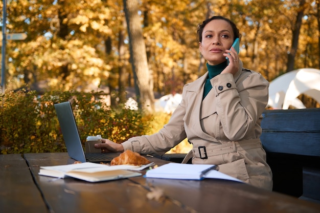 Femme d'affaires confiante regardant sur le côté, parlant au téléphone portable, assise sur un banc en bois devant un ordinateur portable et profitant d'une pause-café dans un café confortable sur fond de chêne d'automne doré