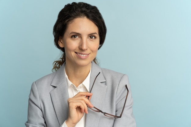 Femme d'affaires confiante tenant des lunettes, souriant, regardant la caméra posant isolé sur fond bleu.