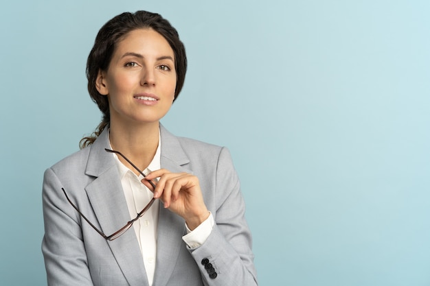 Femme d'affaires confiante tenant des lunettes, souriant, regardant la caméra posant isolé sur fond bleu.