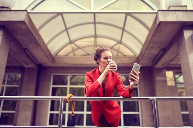 Femme d'affaires en costume rouge à l'aide de téléphone mobile près d'un bâtiment moderne.