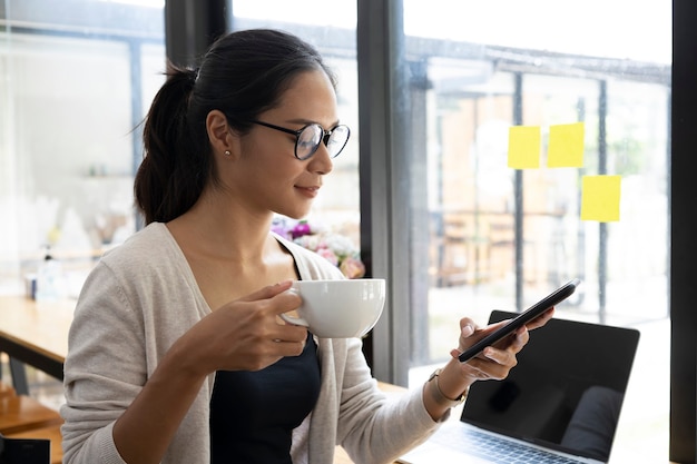 Femme d'affaires dans des verres tenant une tasse de café et utilisant un téléphone portable.