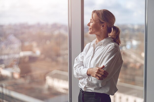 Femme d'affaires debout au bureaumature heureux propriétaire est debout près de la fenêtre du bureau en regardant loin