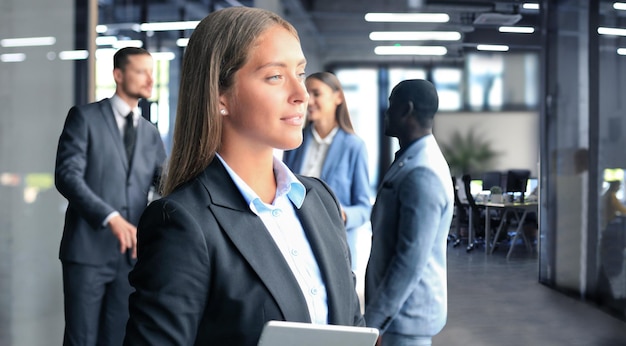 Femme d'affaires debout au premier plan avec une tablette dans les mains, ses collègues discutant de questions commerciales en arrière-plan