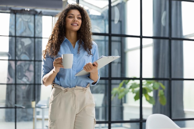 Femme d'affaires debout dans le couloir du bureau avec des documents et du café