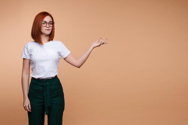 Femme d'affaires décontractée dans des verres montrant la main isolée sur fond de studio. Femme rousse souriante pointant sur place pour le texte posant en regardant la caméra