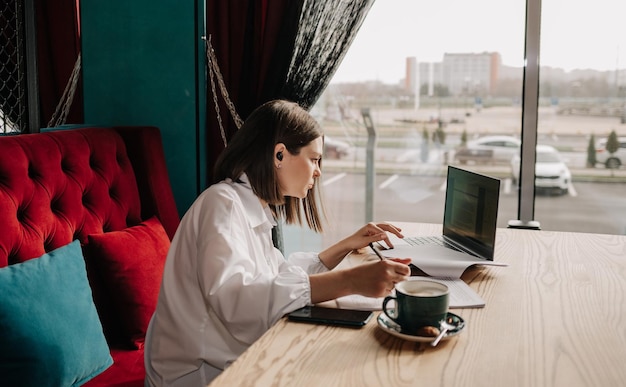 Une femme d'affaires avec des écouteurs sans fil est assise à une table dans un café avec des documents et travaille sur un ordinateur portable