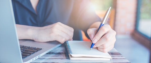 Femme d'affaires écrit à la main sur un bloc-notes avec un stylo et utilise un ordinateur portable dans la bannière web office.for.