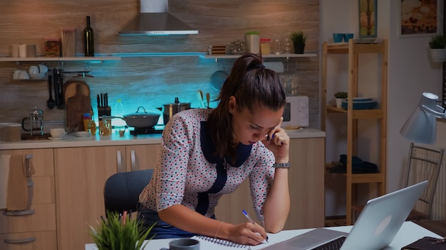 Femme d'affaires écrit des notes tout en travaillant tard sur l'ordinateur dans la cuisine à domicile. Employé concentré occupé utilisant un réseau de technologie moderne sans fil faisant des heures supplémentaires pour la lecture d'emplois, la saisie, la recherche