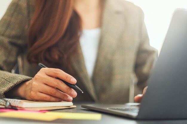 Femme d'affaires écrivant sur de la paperasse tout en tapant sur un ordinateur portable sur la table au bureau