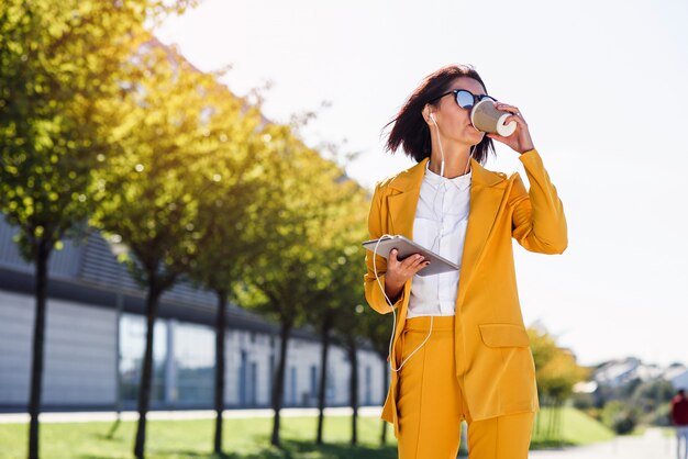 Femme d'affaires élégant en costume jaune avec tablette et casque boit du café tout en se promenant dans la ruelle.