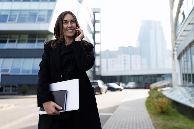 Femme d'affaires élégante avec un ordinateur portable dans ses mains avec un sourire sur son visage parle sur un téléphone mobile