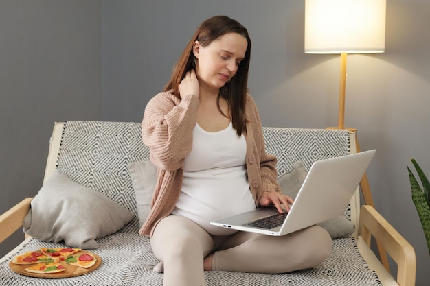 Une femme d'affaires enceinte épuisée, professionnelle, assise sur le canapé, souffrant de douleurs au cou tout en travaillant en ligne sur un ordinateur portable, faisant des grimaces de douleur, s'asseyant dans une posture inconfortable, massant.