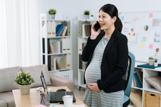 Femme d'affaires enceinte parlant au téléphone portable tout en se tenant près du bureau dans un bureau confortable et lumineux. jeune future maman travailleuse en costume ayant une conversation sur un téléphone portable et une main tenant un gros ventre à l'intérieur