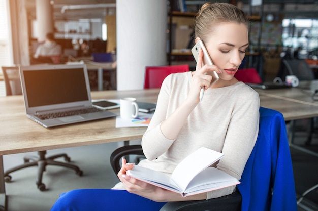 Femme d'affaires est assis sur une chaise et tient un cahier. Elle parle aussi au téléphone. la femme regarde vers la gauche. Elle est concentrée là-dessus.