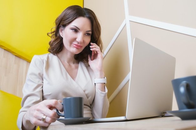 Une femme d'affaires est assise dans un café devant un écran d'ordinateur portable et parle sur un téléphone portable