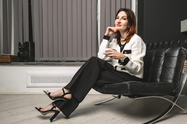 Une femme d'affaires est assise dans une chaise en cuir basse avec une tasse de café dans les mains.