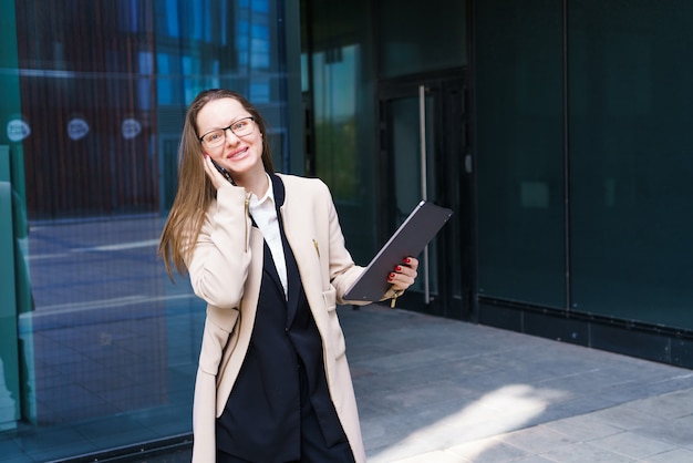 Femme d'affaires ethnique caucasienne en costume et lunettes avec ordinateur portable dans ses mains parlant au téléphone près...