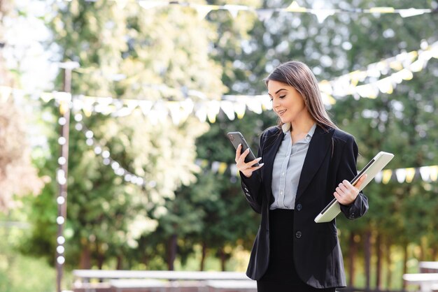 Une femme d'affaires européenne utilise son téléphone portable à l'extérieur Une jeune fille belle et concentrée porte un costume formel Parc ensoleillé pendant la journée Style de vie de la femme moderne Concept de travail à distance et indépendant