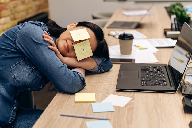 Femme d'affaires fatiguée allongée sur le bureau et dormant avec des autocollants sur les yeux devant l'écran de l'ordinateur. Concept fatigué et surmené