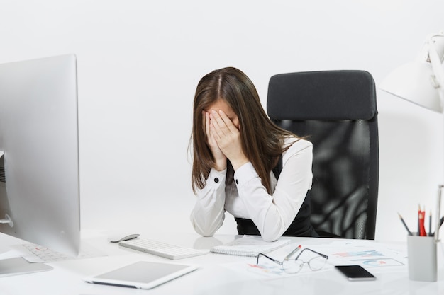 Femme d'affaires fatiguée, perplexe et stressée aux cheveux bruns en costume assise au bureau, se couvrant le visage avec les mains, travaillant sur un ordinateur contemporain avec des documents dans un bureau léger